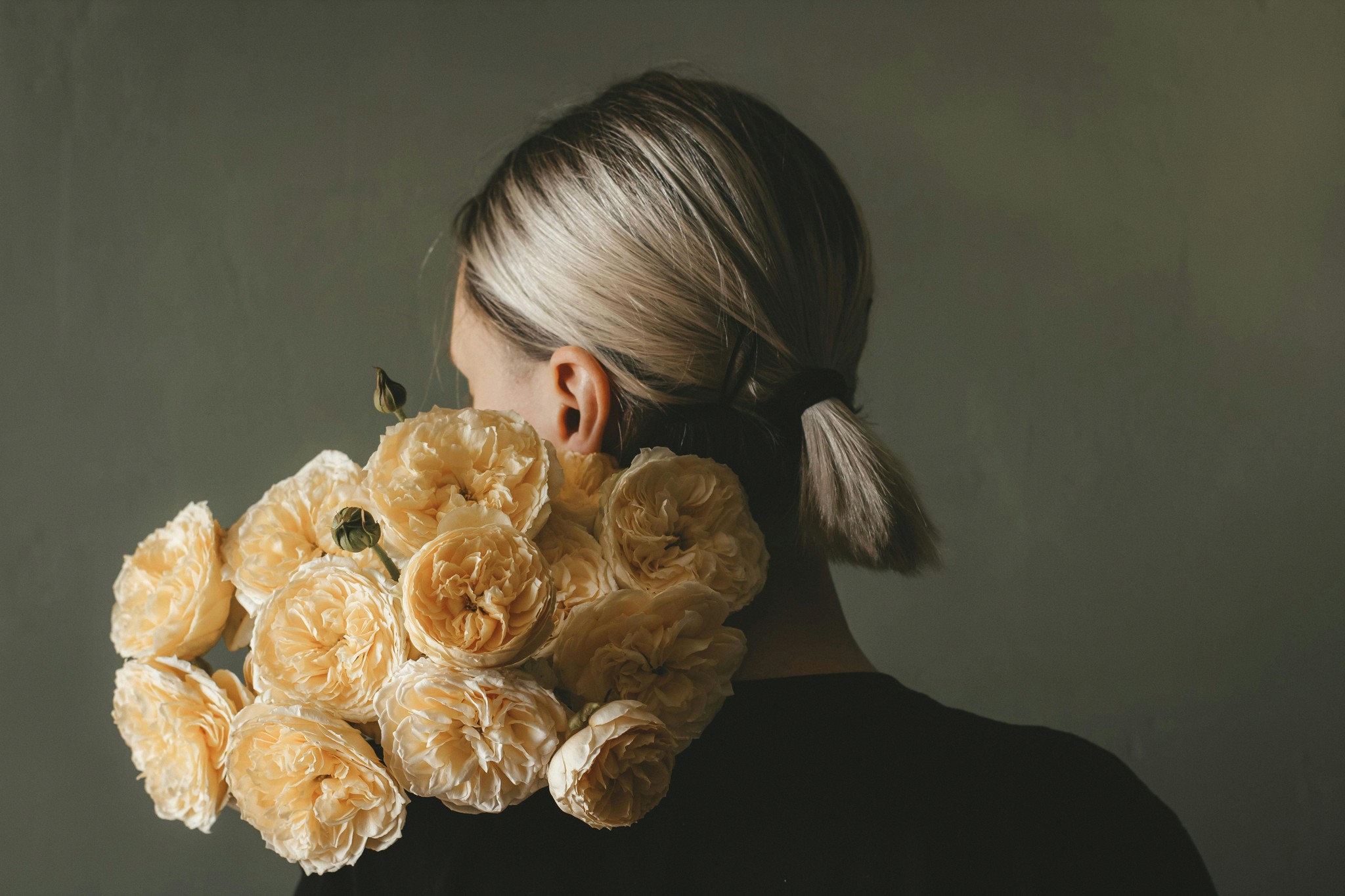A person with blonde hair in a ponytail holding a bouquet of pale yellow roses over their shoulder against a dark wall.